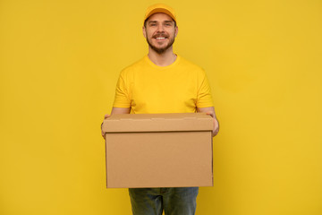 Portrait of excited delivery man in yellow uniform holding paper box isolated over yellow background.