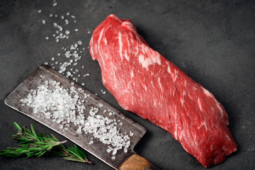 Canvas Print - Raw tenderloin of beef or skirt steak with a hatchet for meat, salt and rosemary on a dark stone background, close up