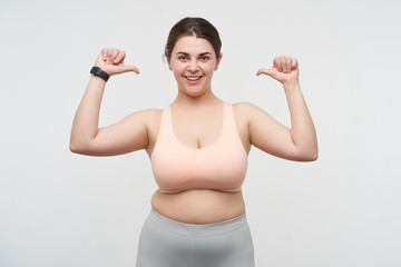 Studio shot of young happy cute dark haired fatty lady with ponytail hairstyle thumbing on herself while looking cheerfully at camera, standing over white background