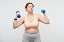 Indoor Photo Of Young Brunette Chubby Lady With Casual Hairstyle Looking Seriously At Camera While Working Out With Dumbbells, Isolated Over White Background