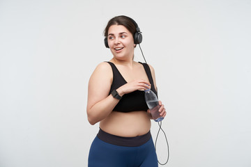 Positive young lovely brunette oversized female drinking water while having fitness class and being in high spirit, posing over white background. Body positive concept