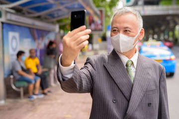 Poster - Mature Japanese businessman with mask taking selfie at the bus stop