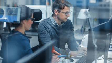 Shot Through Glass in Modern Factory: Female Project Supervisor Talks to a Male Industrial Engineer who Works on Personal Computer. Modern High-Tech Industrial Factory Office.