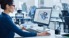 Industrial Female Engineer Working On A Personal Computer, Screen Shows CAD Software With 3D Prototype Of Engine. Busy Factory With Professional Workers High-Tech Machinery