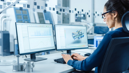 Leinwandbilder - Industrial Female Engineer Working on a Personal Computer, Two Monitor Screens Show CAD Software with 3D Prototype of New Hybrid Hydrogen Engine and Charts. Modern Factory with High-Tech Machinery