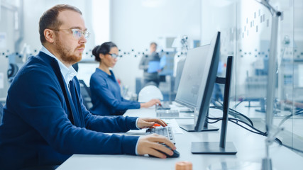 Inside Factory Glass Office Team of Industrial Engineers Working on PC, Outside Cubicle in the Modern Facility Workshop Professional Workers Using High-Tech Industrial CNC Machinery