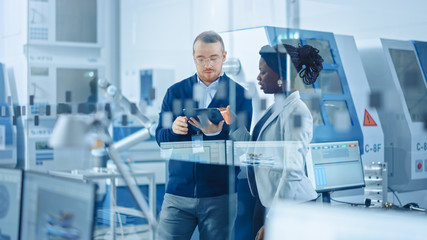 Leinwandbilder - Female Industrial Engineer stands with Male Project Supervisor, They Use Tablet Computer and Discuss Modern Factory Planning. Manufacturing Facility with CNC Machinery and Robot Arm Assembly Line