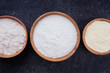 Rice flour, corn flour and whole grain flour in wooden bowls on dark background. Flat lay top view, studio shot