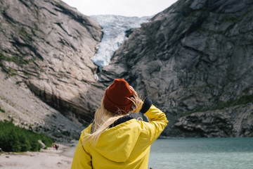 Wall Mural - Blonde woman in yellow raincoat and terracotta hat feels freedom and looks how blue ice tongue of Briksdal glacier slides from the giant rocky mountain and melts into cold lake. Trip to Norway