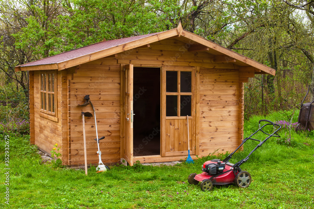 Garden shed with hoe, string trimmer,  rake and grass-cutter. Gardening tools shed. Garden house on lawn in garden. Wooden tool-shed. Hovel made of timber in domestic environment. - obrazy, fototapety, plakaty 