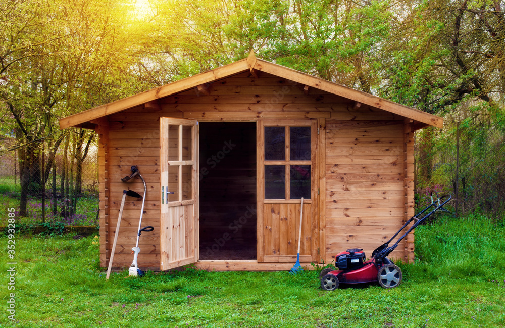 Hovel after work in evening, golden hour. Garden shed (front view) with hoe, string trimmer,  rake and grass-cutter. Gardening tools shed. Garden house on lawn in the sunset. Wooden tool-shed. - obrazy, fototapety, plakaty 