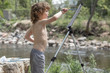 A boy is painting outside in the woods, the kid is shirtless in the summer. He is creative, having fun with art, paint and his imagination in nature. He is by a lake, pond, stream of water. 