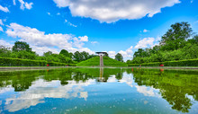 Public Park In Berlin, Germany, In Honor Of The Aviation Pioneer Otto Lilienthal With A Pool Of Water And Reflections Of The Sky In The Foreground And The Monument On A Hill In The Background.