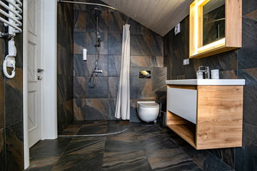 Interior of modern stylish bathroom with black tiled walls, curtain shower place and wooden furniture with wash basin and big illuminated mirror.