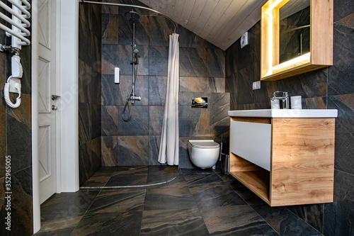 Fotografía Interior of modern stylish bathroom with black tiled walls, curtain shower place and wooden furniture with wash basin and big illuminated mirror