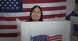 © vesperstock - Front view MS mature Caucasian woman looks up, thoughtfully considering, while voting in booth at polling station. Large US flag on wall behind.