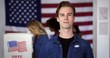 © vesperstock - MCU Young Caucasian man in denim shirt with 'I Voted' sticker, smiles, standing proud in front of other voters at polling booths with US flag behind