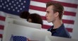 © vesperstock - Close up profile, young Caucasian man looking ahead, thoughtfully, while casting vote at polling station during election. Other voters in background and large US flag on wall behind.