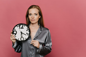 Beautiful confident redhead caucasian female in nightwear, holds wall clock, looks calm, always be in time, waking up early, standing pink background with copy space.