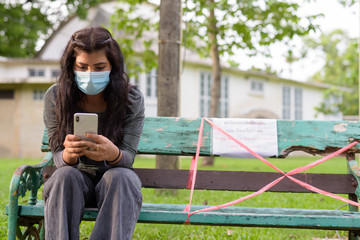 Poster - Young Indian woman with mask using phone while sitting with distance on park bench