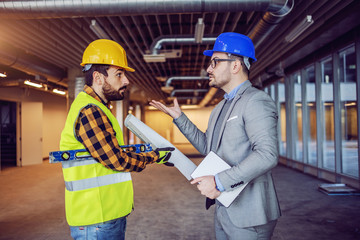 Angry caucasian businessman in suit and helmet on head arguing with irresponsible construction worker. Building in construction process interior.