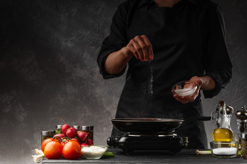 The chef is fried in a pan and salted. Freeze in motion. Against the background of vegetables. Culinary recipes and home cooking.