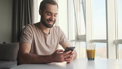 Canvas Print - A smiling positive young man is watching something funny on his smartphone sitting at the table at home 