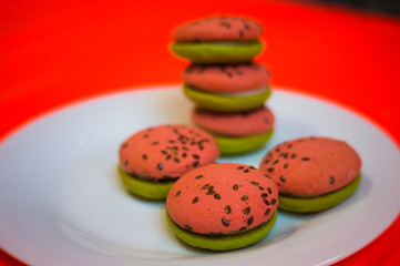 Multi-colored green and pink, bright cookies with two halves dressed with filling and sprinkled with sesame seeds on a white plate and on a bright pink background.