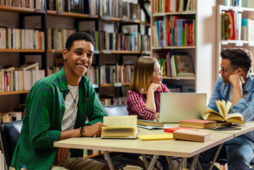 Wall Mural - Portrait of young african american student sitting in library looking at camera, with his friends in background.