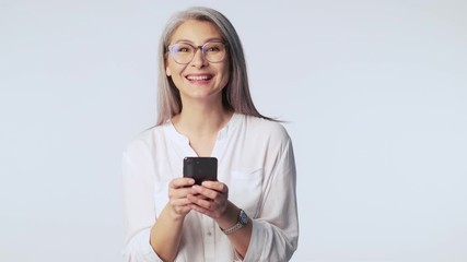 Wall Mural - A happy pleased old mature woman with long gray hair wearing glasses is typing on her smartphone standing isolated over white background in studio