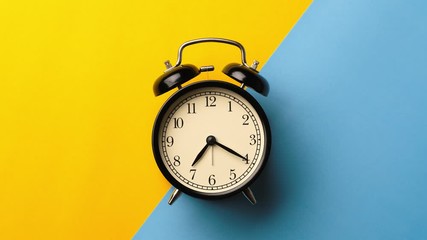 Wall Mural - Time on black vintage alarm clock running from 7:00 to 8:00 isolated on two tone color yellow and light blue background color. One hour time lapse of black alarm clock running from 7:00 to 8:00.