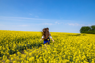 Wall Mural - young woman running in a field with yellow flowers, blue sky, space, girl in an embroidered shirt and blue skirt, long hair