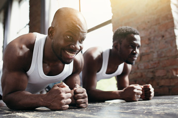 Two African-Americans in white T-shirts train the abs and back muscles while standing with their forearms and legs on the floor.