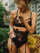 Young beautiful girl in black lingerie and a beige trench coat drinks champagne on a balcony in the morning