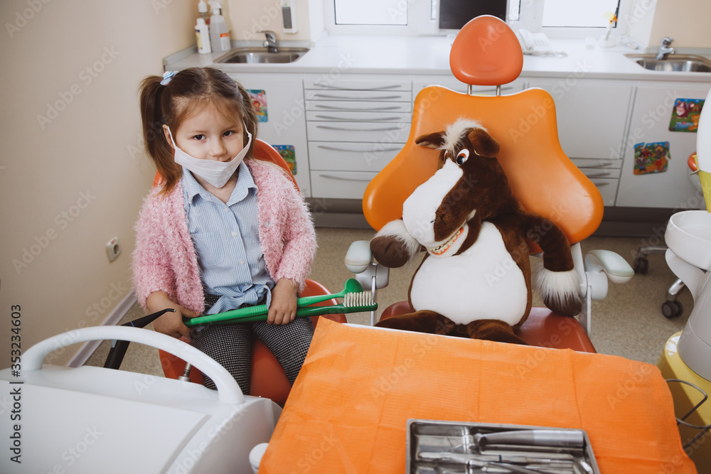 High angle of little girl in medical mask sitting near chair with toy ...