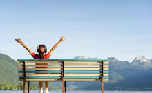 Girl Sitting On A Bench With Her Arms Up With Stalk Hat On And Red Dress Relaxing Looking At The Mountains By A Lake