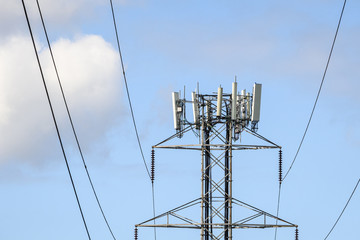 High transmission power lines, self-supporting tower with mobility antennas on top, late afternoon blue sky with white clouds in background
