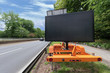 © betzalit - a blank digital highway shoulder sign, for copy space, with the road on one side and a car driving away in the distance, surrounded by foliage. A blue sky with wispy clouds in the background.