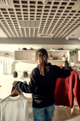 Poster - Buy local. Young woman holding hangers with custom apparel on it, standing in the store. Clothes at custom T-shirt, clothing printing company