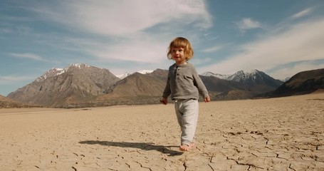 Wall Mural - Funny caucasian baby boy running on deserted ground. Cracked soil land after erosions and desertification - ecology, save our planet concept 4k footage