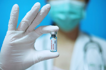 A female Asian physician with surgical mask and white rubber gloves at a clinic, holding a glass bottle of 1 dose PCV vaccine with white background and red letters.