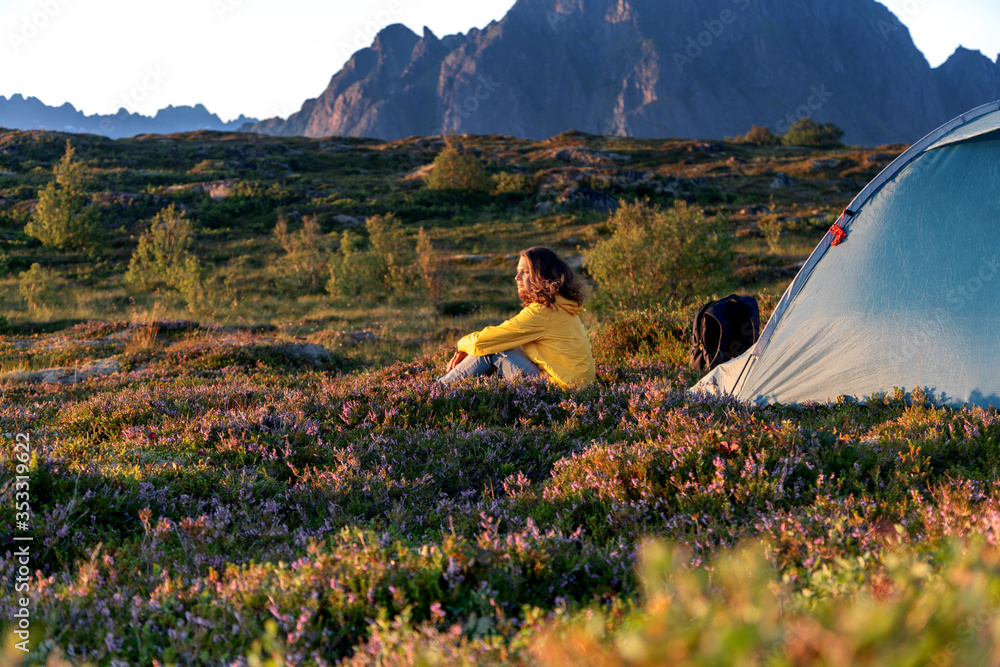 A young woman in yellow  jacket sits next to a camping tent on the background of a beautiful landscape at sunset, traveling to Norway Lofoten islands - obrazy, fototapety, plakaty 