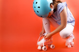 Little child with roller skates, a blue helmet on a red background, tying shoelaces. A girl of 7 years old poses and prepares for active leisure on retro ice skates. Copy space