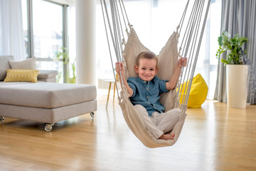 Adorable little smiling boy enjoying on indoor swing in bright modern living room