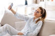 Selfie on the phone. Young beautiful girl with attractive appearance of caucasian nationality takes a selfie on the phone while sitting on a sofa at home in casual stylish clothes