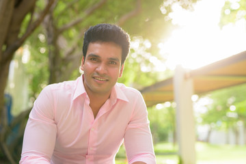 Happy young handsome Indian businessman smiling at the park outdoors