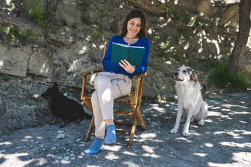 Young smiling woman sitting on a chair in the garden reading a book with two dogs around. Open air activities and relax concept.