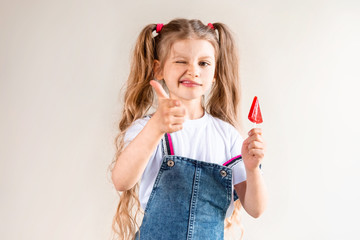 A little girl with ponytails holds a red Lollipop on a stick and aims with her index finger.