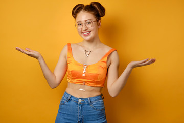 The girl with two beams, glasses and an orange top stands on a yellow background and smiles, spreading her palms to the sides