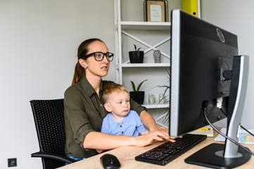 Wall Mural - Modern busy mother working with a baby in the home office. A young woman typing on pc keyboard while cute boy kid sits on her laps